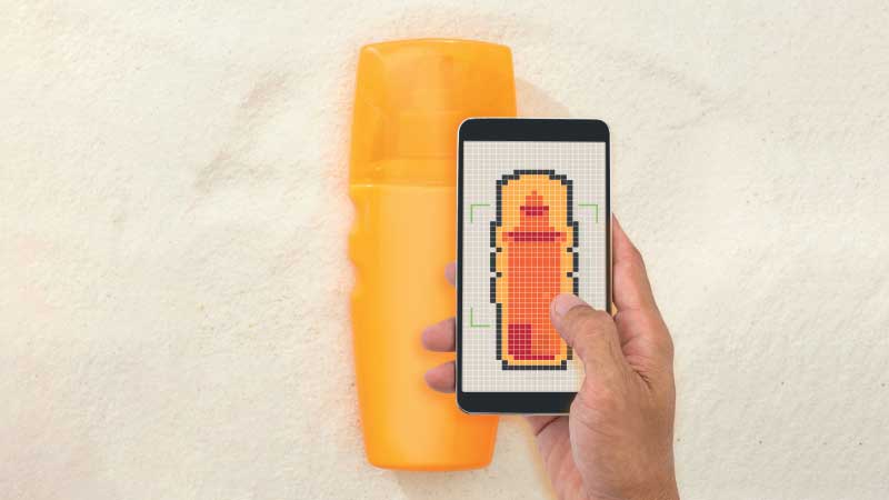 Mão segura telemóvel apontado a uma embalagem amarela de protetor solar, para verificação do conteúdo, como se fosse um scanner